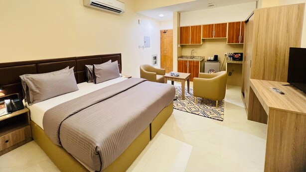 Habitación de hotel con cama, mesita de noche, lámpara y un rincón de cocina equipado con lavabo y armarios.