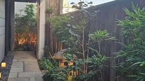 Un étroit sentier de pierres mène à une construction en bois surmontée d’un toit de chaume, entourée de plantes de bambou et d’une clôture en bambou.
