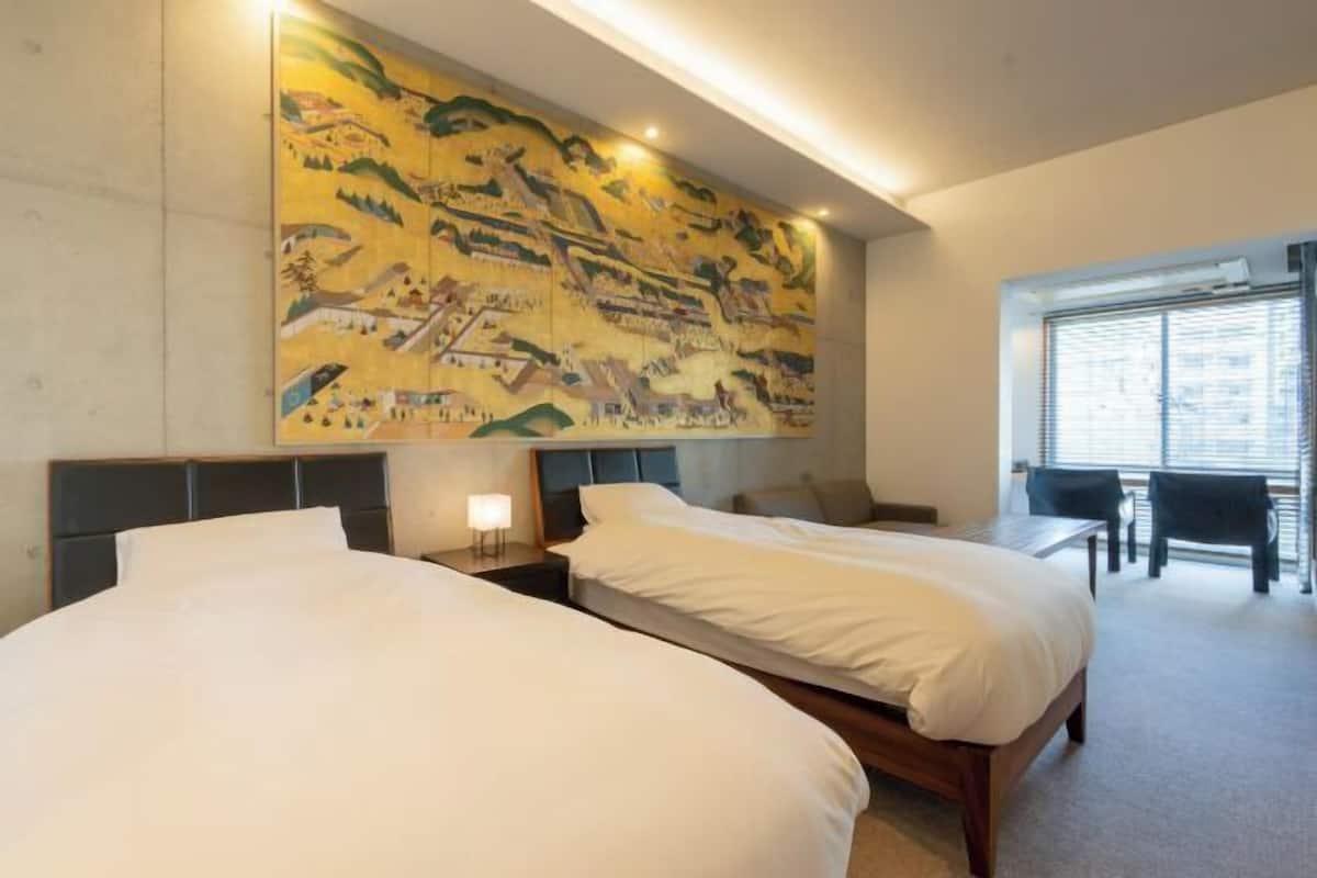 Habitación de hotel con dos camas, un mural grande en la pared, una mesita y una ventana con persianas.