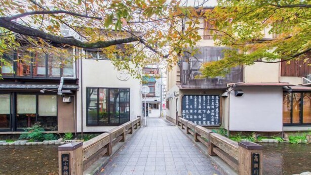 Un sentier mène à un bâtiment traditionnel, et un pont en bois enjambe un bassin d’eau ou un autre élément aquatique.