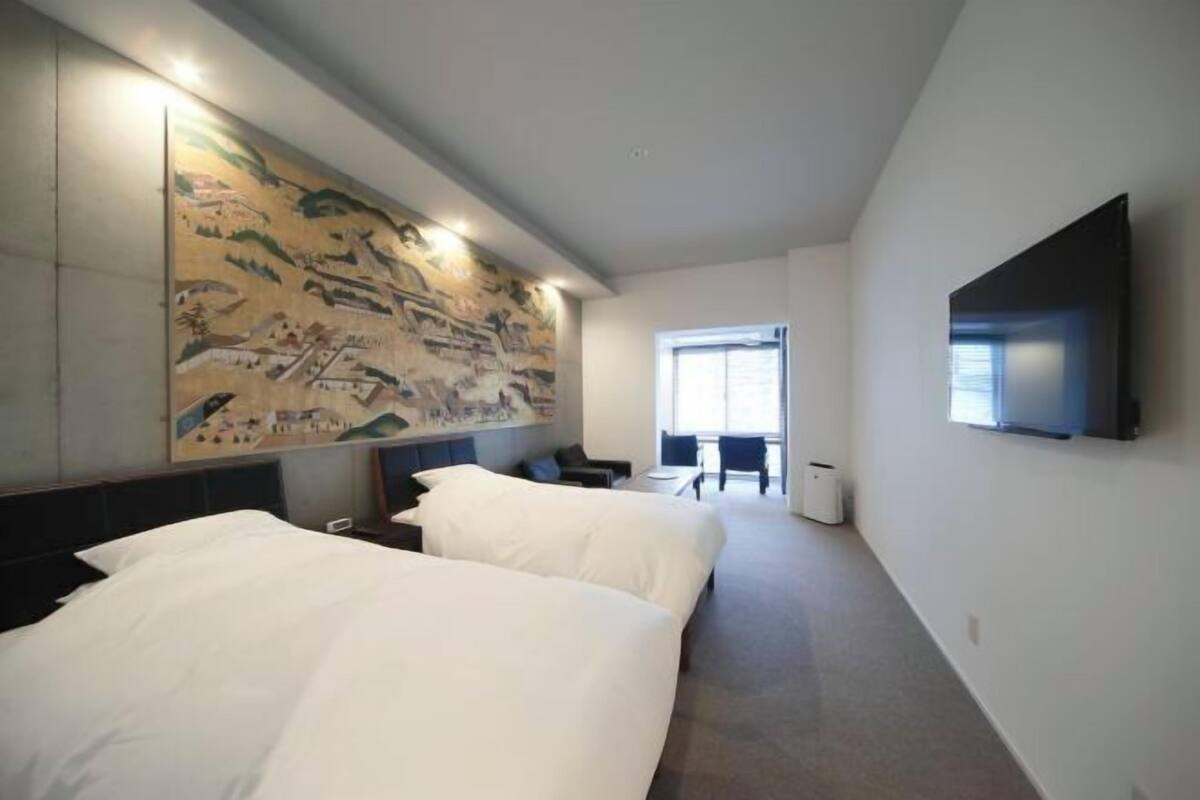 Habitación de hotel con dos camas, un mural grande en la pared, un televisor de pantalla plana montado y una ventana que proporciona luz natural.