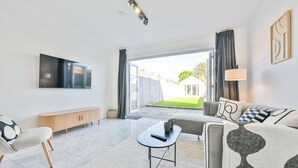 A modern living room with a flat-screen TV, a sofa, a coffee table, and a view of a garden through a sliding glass door. - The Grove by Tŷ SA (Cardiff)