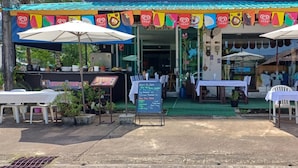 An outdoor dining area with tables and umbrellas, a menu board, and a colorful awning. - Burmese Corner (Khao Lak)
