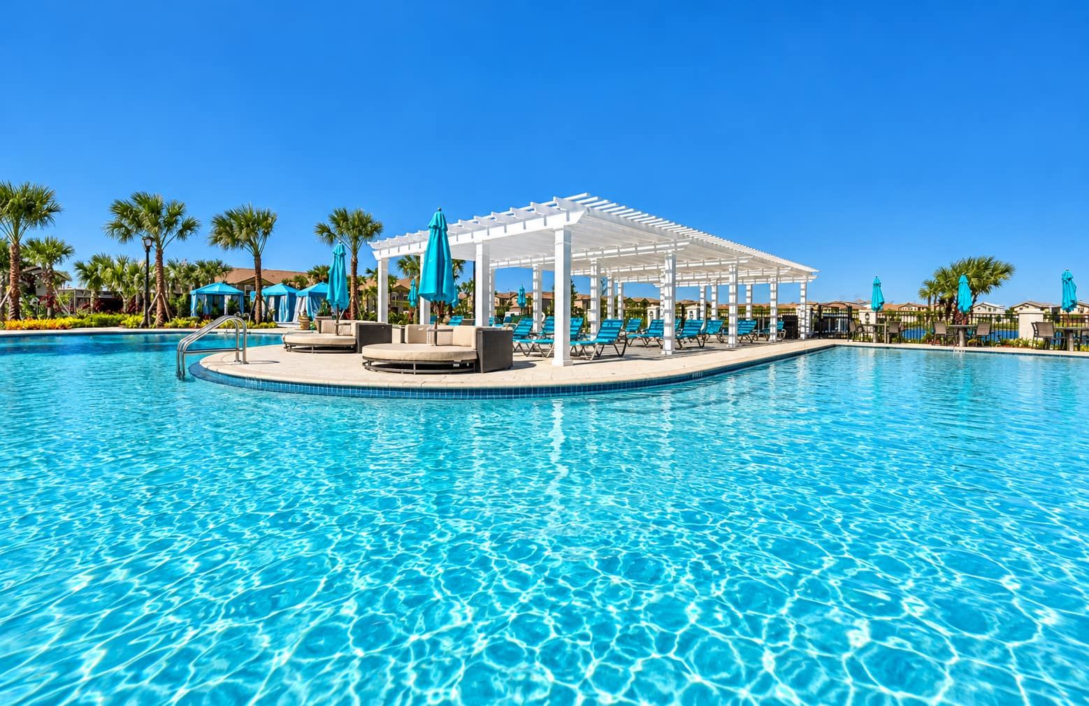 A swimming pool with lounge chairs and umbrellas, surrounded by palm trees and a covered seating area.