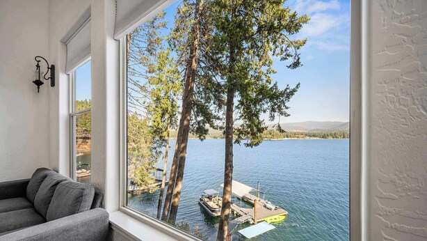 A room with a large window overlooking a lake, a gray sofa, and a small table. - Lakefront Hayden Lake Home with Private Dock for Family Getaways (Hayden)