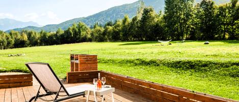Eine Holzterrasse mit Liegestuhl und Tisch, mit Blick auf eine Wiese und Berge.