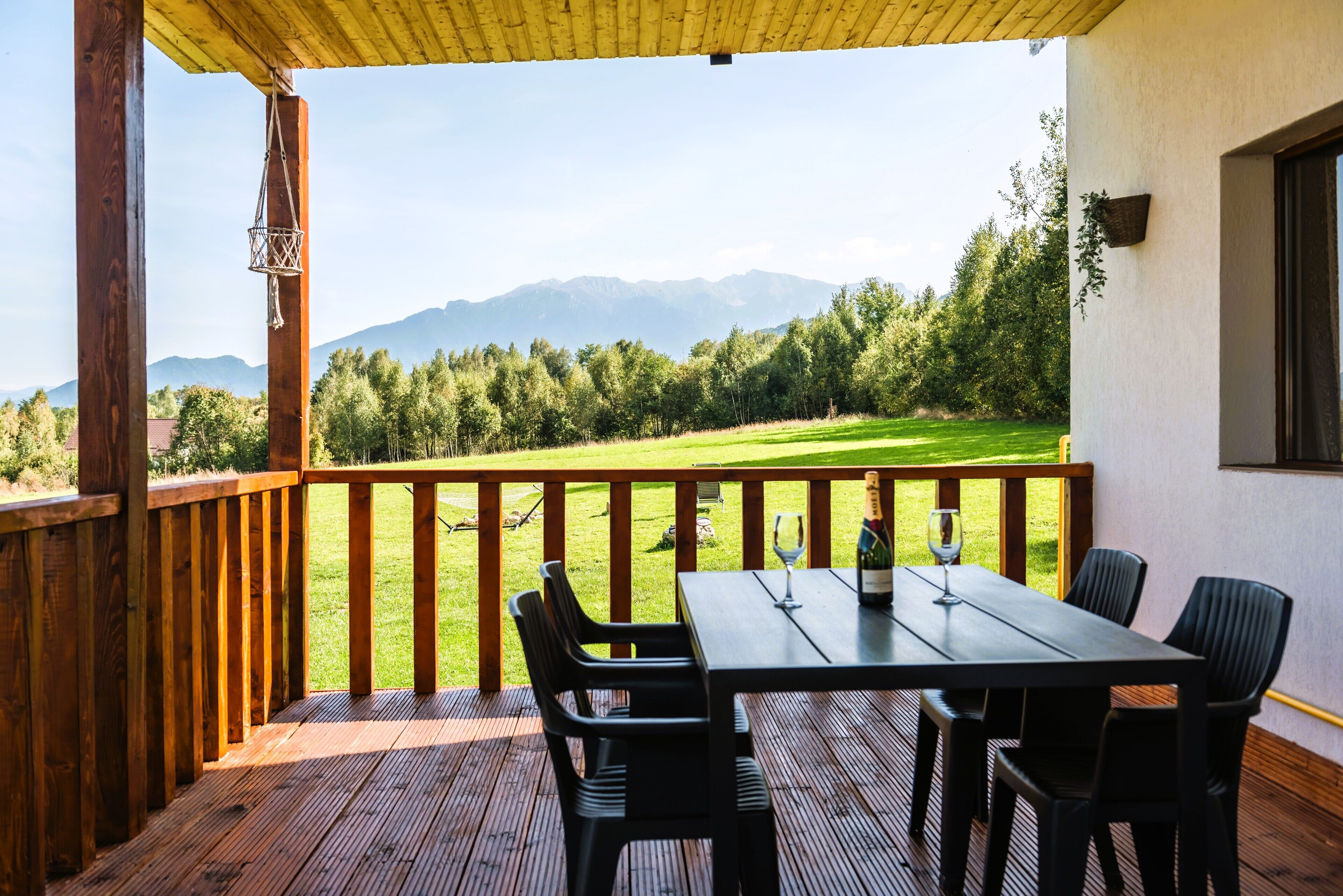 A wooden deck with a table and chairs, overlooking a grassy area and mountains.