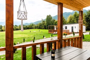 A wooden deck with a bottle of champagne, overlooking a grassy area with a small building and mountains in the background. - RAI Lodges (Zărnești)