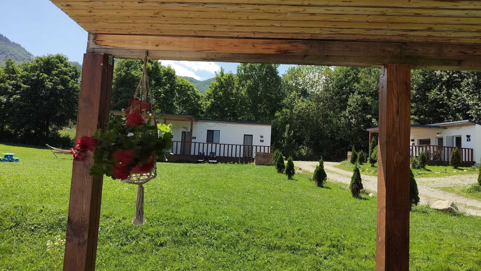 Une structure en bois formant une pergola, agrémentée d’une décoration suspendue, avec une pelouse verdoyante et, au fond, une caravane blanche.