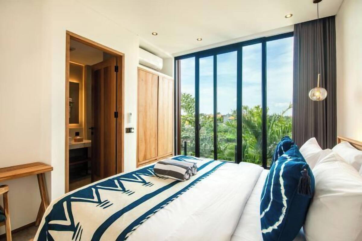 A bedroom with a large window, a bed with patterned bedding, a wooden wardrobe, and a small table.