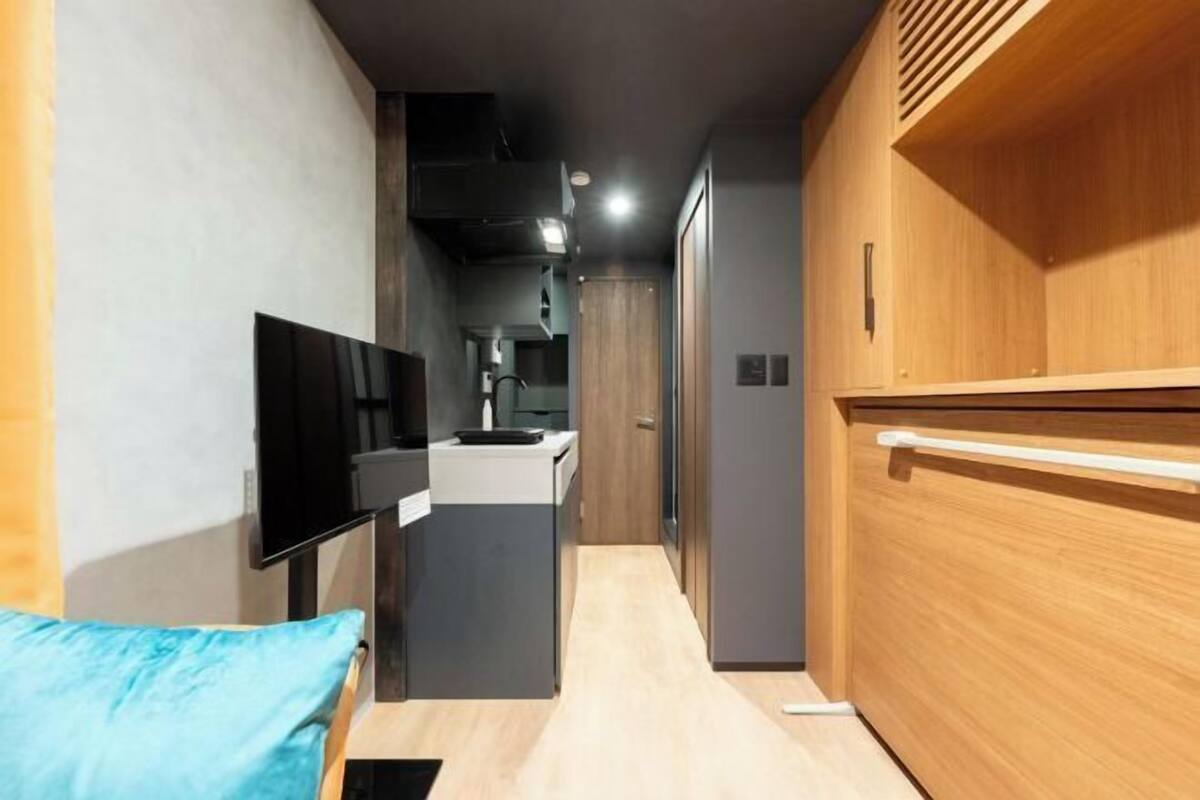 A narrow hallway leading to a kitchen area with a TV mounted on the wall, a wooden cabinet, and a light-colored floor.