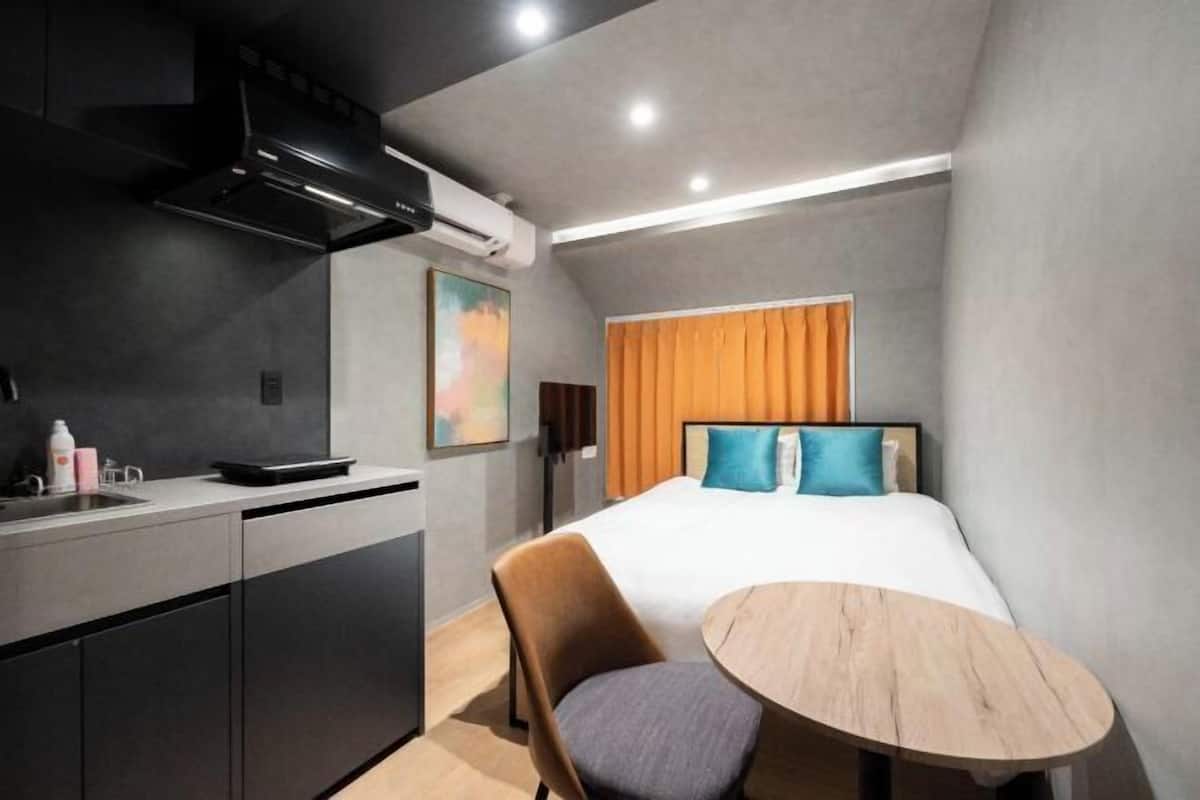 A compact hotel room with a kitchenette, a bed with blue and teal pillows, a round wooden table, and a window with orange curtains.