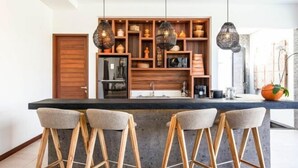 A modern kitchen with a central island, wooden bar stools, pendant lighting, and a variety of kitchenware on shelves.
