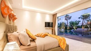 A modern bedroom with a bed, a TV, and a view of a pool and palm trees.