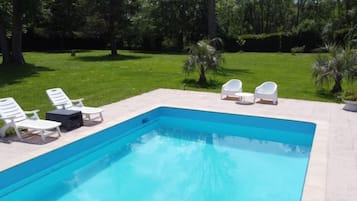 Uma piscina retangular com superfície de água azul cristalina, rodeada por um deck pavimentado. Quatro cadeiras de espreguiçadeira brancas estão alinhadas na borda da piscina.