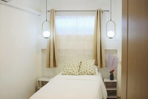 A bedroom with a bed, two bedside tables, a nightstand, a window with curtains, and a hanging light fixture.