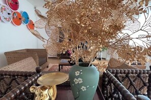 Una habitación con un sofá de mimbre, un jarrón verde con flores blancas y un objeto dorado decorativo sobre una mesa.