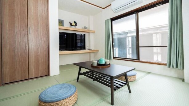 A room with tatami flooring, a low table with a cushion, a wardrobe, a shelf with decorative items, and a large window with curtains. - Kaike・Honcho detached house (Tokyo)