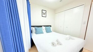 Een opgemaakt bed met witte beddengoed en blauwe details, een lijst met een vlinder afbeelding aan de muur, en een rij witte kasten aan de rechterkant.