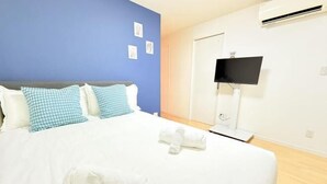 A hotel room with a bed, blue and white checkered pillows, a white blanket, and a TV mounted on the wall. - BASE36 (Tokyo)