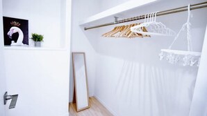 Een witte kamer met een houten vloer, een hangende houten kapstok met kleding, een ingelijst beeld aan de muur en een kleine plant in een pot.