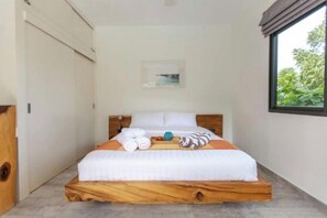 A single bed with a wooden frame and white bedding, a window with a view of greenery, and a door to the left. - Villa Lotus, 3 Bedrooms (Koh Samui)
