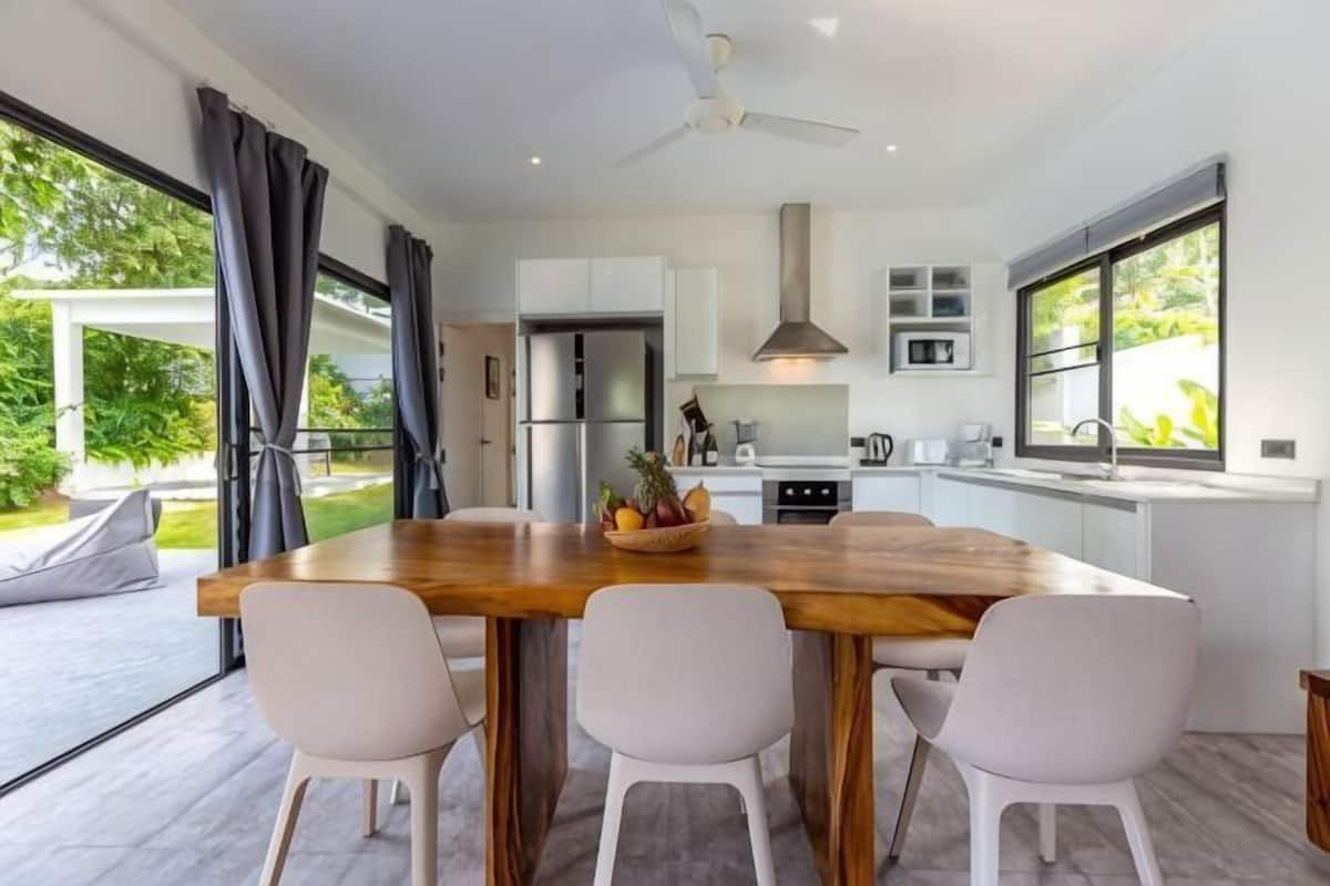A modern kitchen with a wooden island, white chairs, and a view of the outdoors.