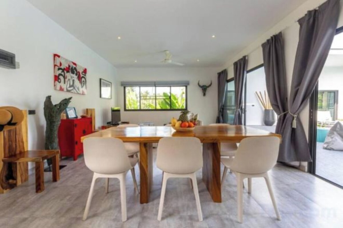 Un coin repas avec une table en bois et quatre chaises, un buffet rouge et une grande fenêtre avec des rideaux.