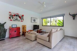 A modern living room with a sofa, a coffee table, and a TV. There is a window with a view of greenery outside. - Villa Lotus, 3 Bedrooms (Koh Samui)