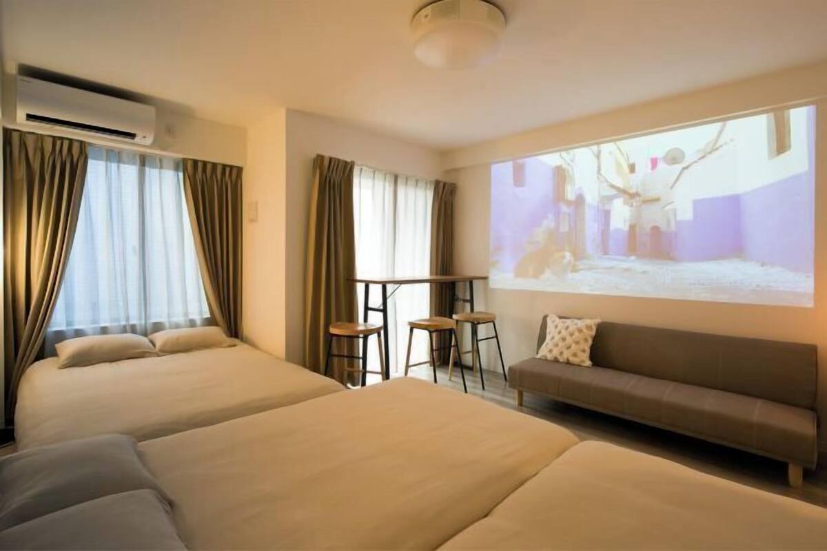 Une chambre d’hôtel avec deux lits, un canapé, une petite table avec des tabourets et une grande fenêtre avec des rideaux.