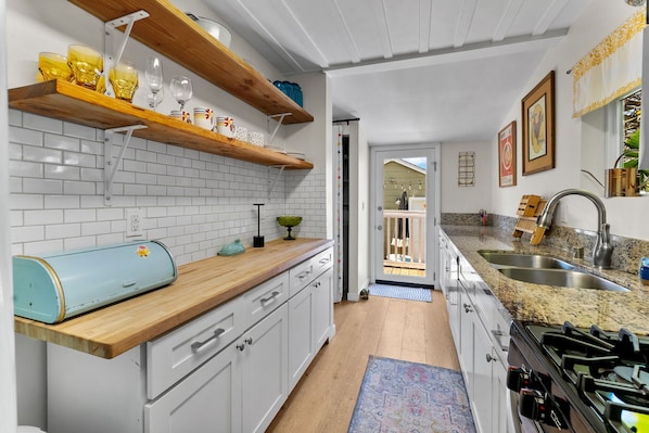 Una cocina con armarios blancos, encimeras de madera y una estufa. Se observan estantes con cristalería y un electrodoméstico azul en la encimera.