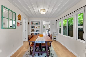 Un comedor con una mesa de madera, sillas y un tapete estampado. Se observa una estantería con libros y objetos decorativos.