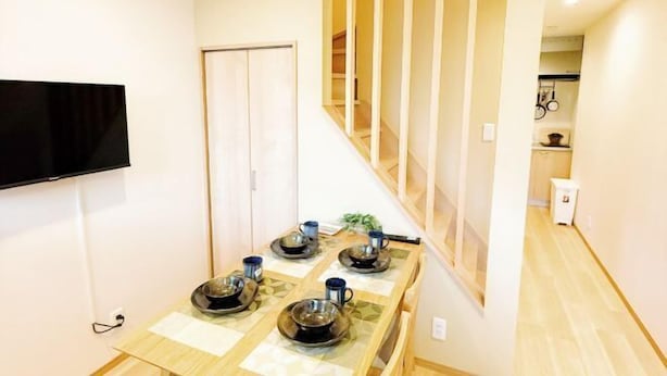 A dining area with a wooden table set for four, a TV mounted on the wall, and a staircase leading to another level. - KIN'S HOUSE (Osaka)