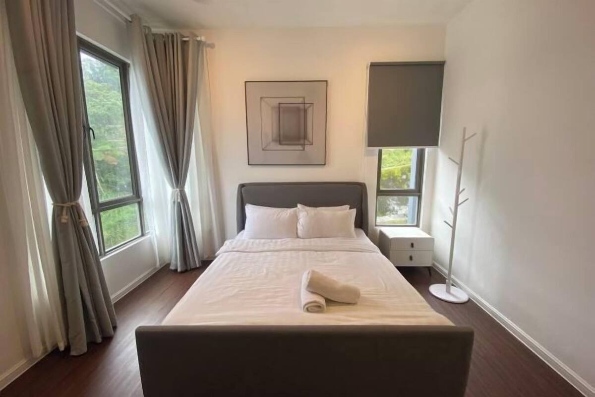 Een opgemaakt bed met een opgerolde handdoek, een nachtkastje, een lamp en een raam met gordijnen.