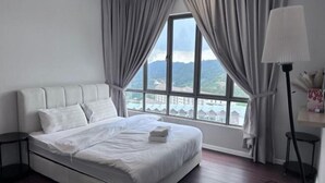 Una cama bien hecha con ropa blanca, una mesita de noche con lámpara, y vistas a un paisaje montañoso a través de un ventanal con cortinas.