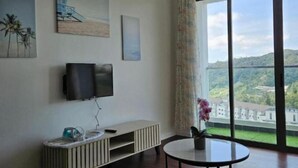 Una habitación con televisión montada en la pared, una mesita con un florero y un ventanal con vistas a zonas verdes y edificios.