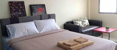 A neatly made bed with folded towels, a sofa, a small table with a plant, and artwork on the wall.