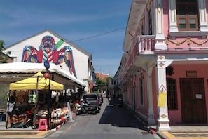 A narrow street with buildings on either side, a market stall on the left, and a parked car in the middle. - Phuket Town Budget House - Aircon Room #1 (Phuket)