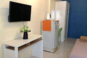 Una habitación compacta con un televisor de pantalla plana montado en la pared, una mesita blanca con una planta en maceta, un refrigerador blanco con una tetera encima y una cama con una manta rosa.