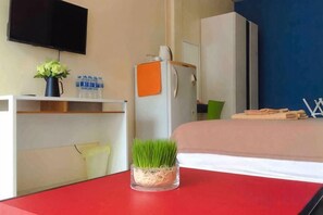 A hotel room with a bed, a desk with a vase of flowers, a television on the wall, and a refrigerator. - Phuket Town Budget House - Aircon Room #1 (Phuket)