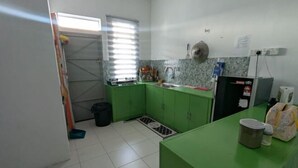 A kitchen with green cabinets, a sink, a window with blinds, a trash bin, and a refrigerator. - Homestay ibunda (Sabak Bernam)