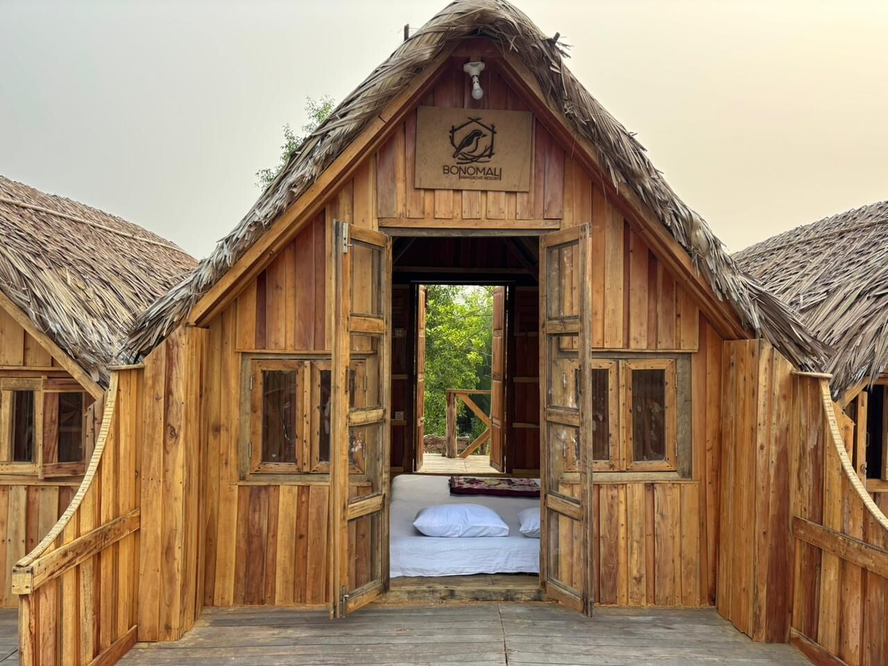 A wooden structure with a thatched roof, an open doorway leading to a bed with white linens, and a logo above the entrance.