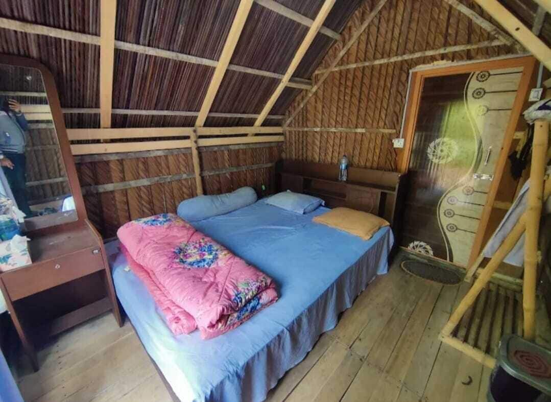 Una cama individual con una colcha azul y una manta rosa, una mesita de noche de madera, un espejo y una puerta con pomo decorativo.