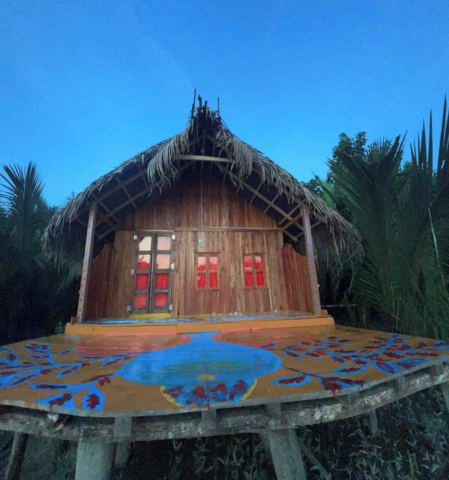 A wooden house with a thatched roof, large windows, and a colorful floor design.