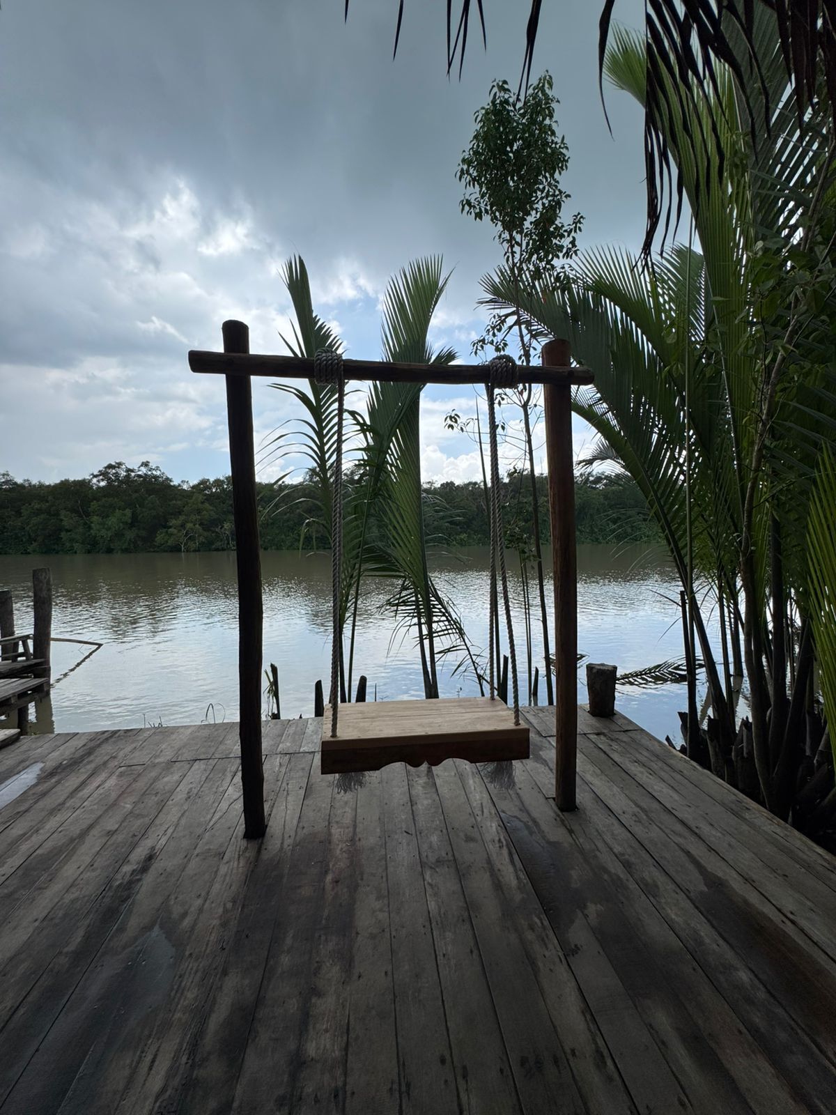 A wooden deck with a swing, surrounded by palm trees and a body of water.