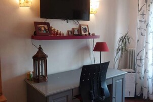 Una habitación con escritorio, silla, televisor montado en la pared, una lámpara roja y una planta.