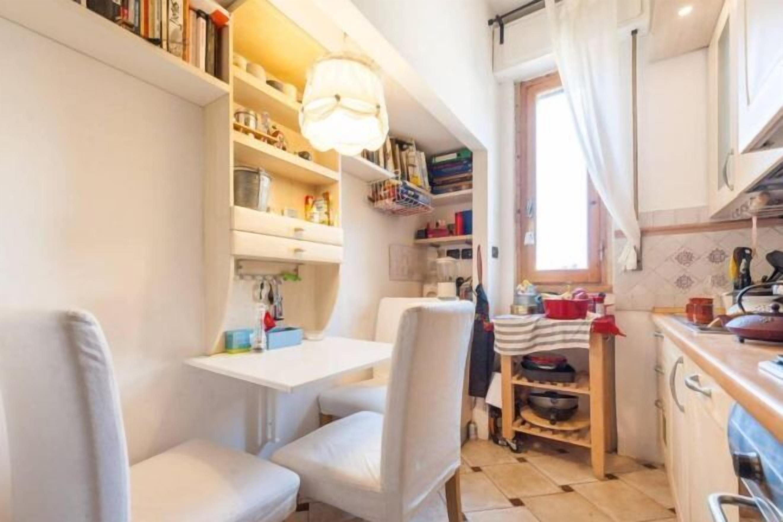 A compact kitchen with white cabinetry, a dining table, and chairs. There are shelves with books and kitchen items, and a window with curtains.