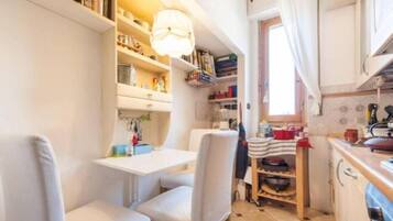 A compact kitchen with white cabinetry, a dining table, and chairs. There are shelves with books and kitchen items, and a window with curtains.