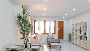 Un comedor moderno con una mesa preparada para cuatro personas, una planta en maceta y una estantería blanca al fondo.