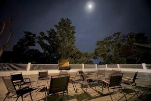 Un espacio al aire libre con sillas y mesas, una cerca blanca y árboles bajo un cielo nocturno con luna visible.